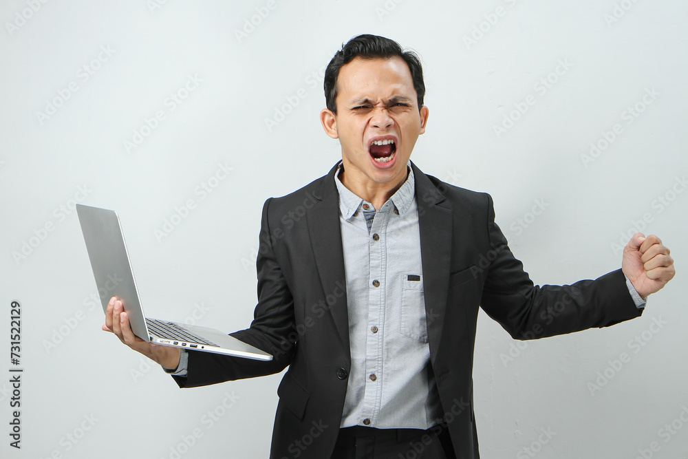 Angry mad asian indonesian business man in suit holding laptop computer on isolated background Angry mad asian indonesian business man in suit holding laptop computer on isolated background