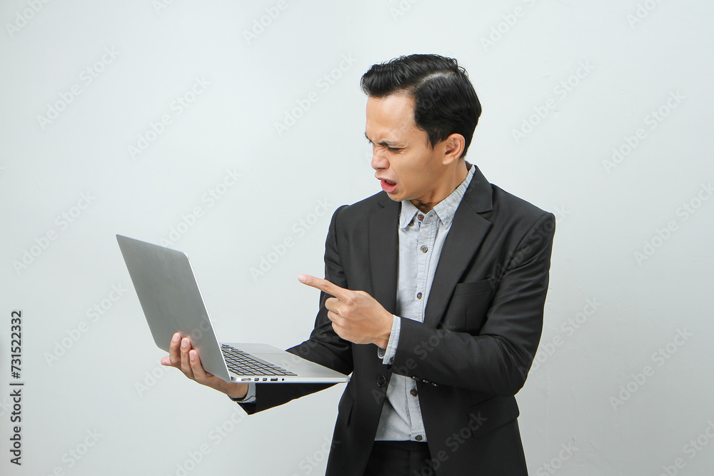 Angry mad asian indonesian business man in suit holding laptop computer on isolated background Angry mad asian indonesian business man in suit holding laptop computer on isolated background