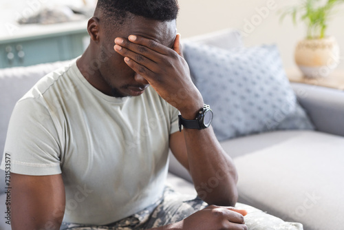 Troubled african american male soldier sitting on couch at home, holding head, copy space photo