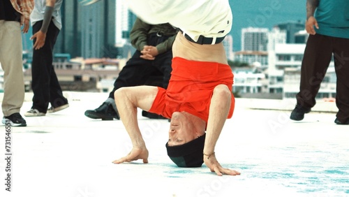 Professional break dancer doing head spin at roof top with city view. Group of skilled caucasian hipster practicing break dancing with multicultural friends encourage behind. Endeavor.