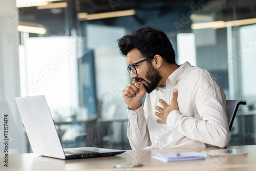 A bearded white-collar worker with eyewear coughing at desk with laptop.