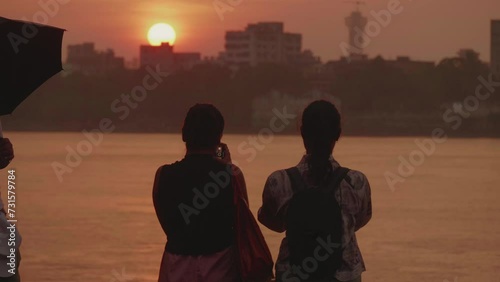 A safety volunteer standing and holding an umbrella watching two girls standing at the Hooghly River or Ganges riverbank clicking pictures of the beautiful sunset during monsoon or rainy season photo
