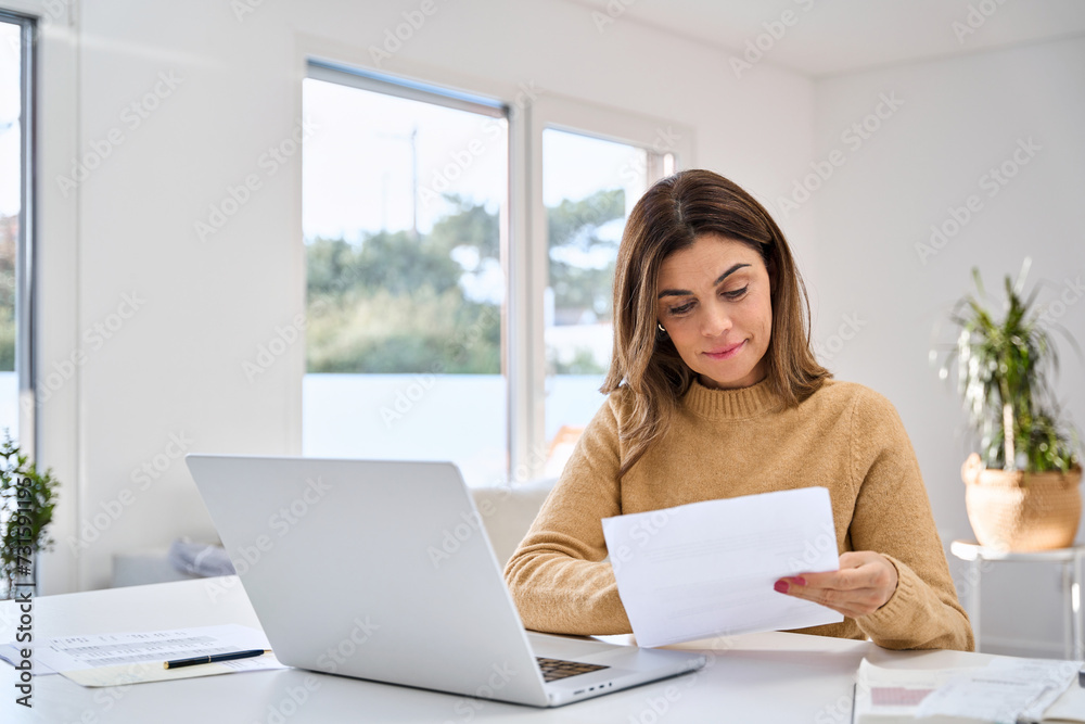 Middle aged older woman reading paper bill working at home using laptop computer in living room. Mature lady checking financial invoice or tax document making online payments working at home. Middle aged older woman reading paper bill working at home using laptop computer in living room. Mature lady checking financial invoice or tax document making online payments working at home.