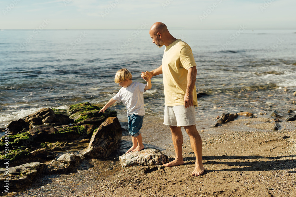 father supports son on rocks in the sea. Shot of father helping his son to jump over the rock on the beach. Young man and little boy having fun at the rocky beach. father supports son on rocks in the sea. Shot of father helping his son to jump over the rock on the beach. Young man and little boy having fun at the rocky beach.