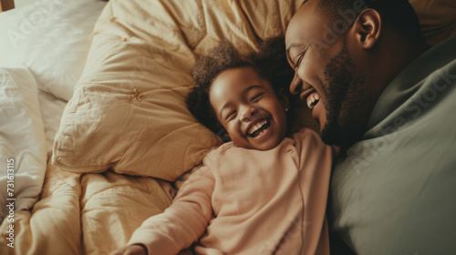 A joyful African American father and his young daughter share a heartfelt laugh while lying together in a cozy, sunlit bed