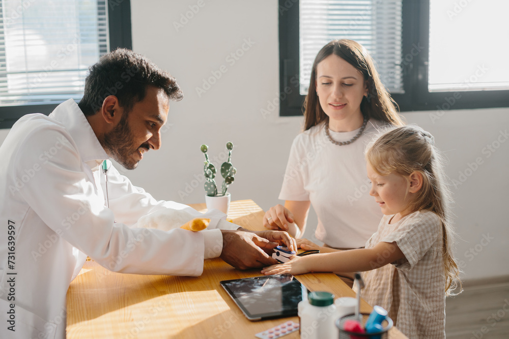 A young pediatrician measures the pulse of a small patient. A mother with a preschool-aged daughter at a doctor's appointment. A young pediatrician measures the pulse of a small patient. A mother with a preschool-aged daughter at a doctor's appointment.