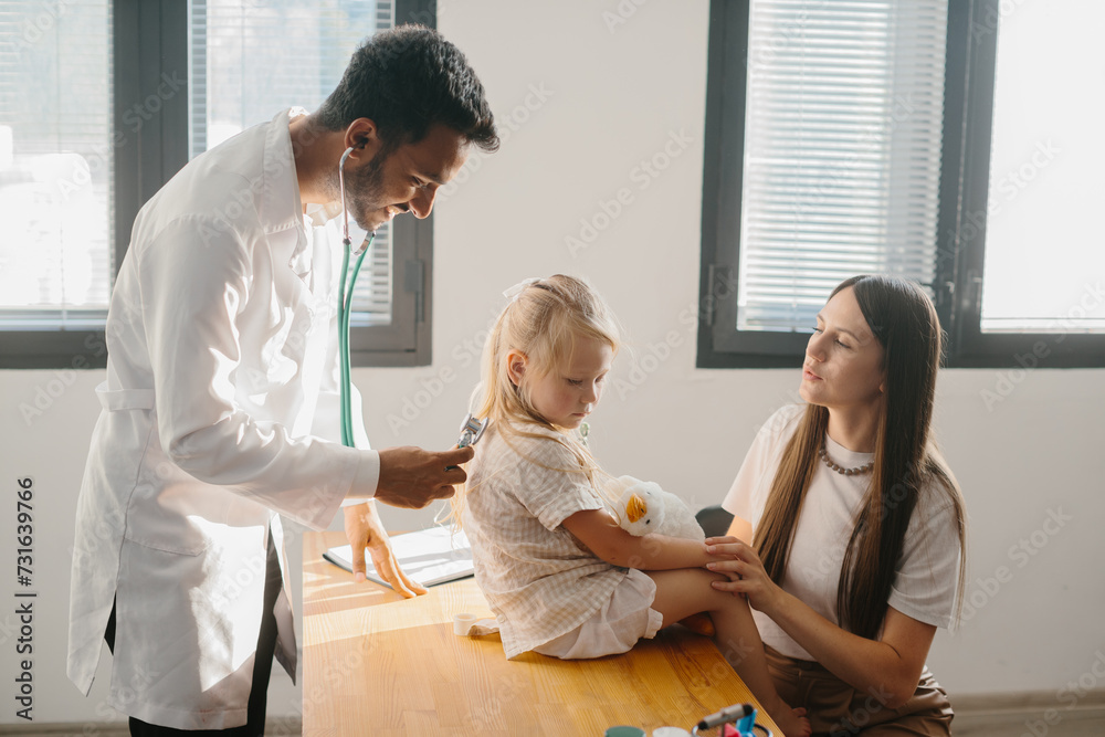 Fototapeta premium A preschool-aged girl with her mother is being examined by a pediatrician. A young man, a pulmonologist, conducts an examination using a phalendoscope.