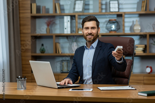 Mature businessman smiling, managing work from home with a laptop and remote control in a well-organized office environment.