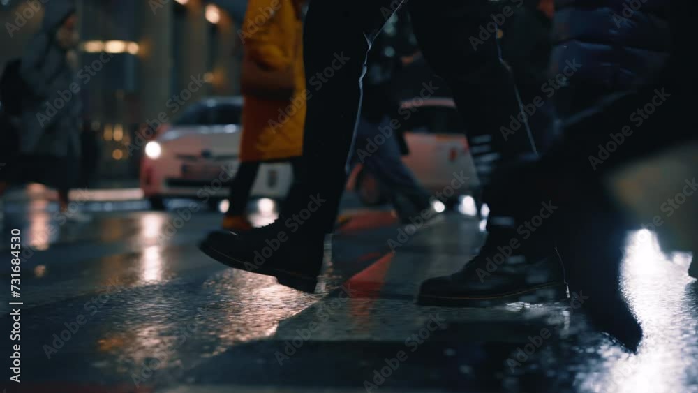 Side view following male feet steps crossing busy street at night. Man wearing leather boots goes on pedestrian crossing city road. People crowd legs walking sidewalk, rainy weather, car lights Side view following male feet steps crossing busy street at night. Man wearing leather boots goes on pedestrian crossing city road. People crowd legs walking sidewalk, rainy weather, car lights