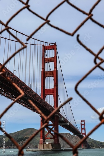 A vertical shot of the Golden Gate Bridge visible through a rusty fence at Fort Point