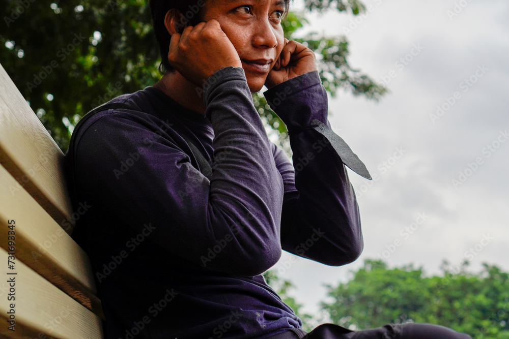 A Javanese young man was sitting on a bench holding his ear as a sign of apology. A Javanese young man was sitting on a bench holding his ear as a sign of apology.