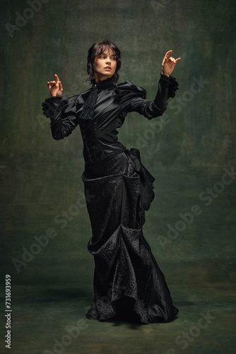 Woman in Victorian style black dress, standing in a dramatic pose against dark vintage green textured background. Concept of retro and vintage fashion, history, baroque style, inspiration