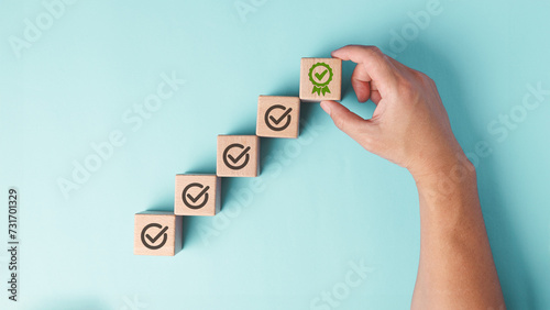 A hand is placing the final wooden block with a green certificate checkmark, symbolizing achievement, quality assurance and completion, against a plain pastel blue background. photo