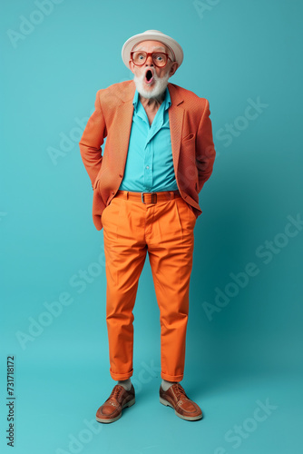 Photo portrait of senior man in blue and orange suit with amazed or surprised expression wearing orange hat. Grandpa is isolated on bright blue color background with copy space.