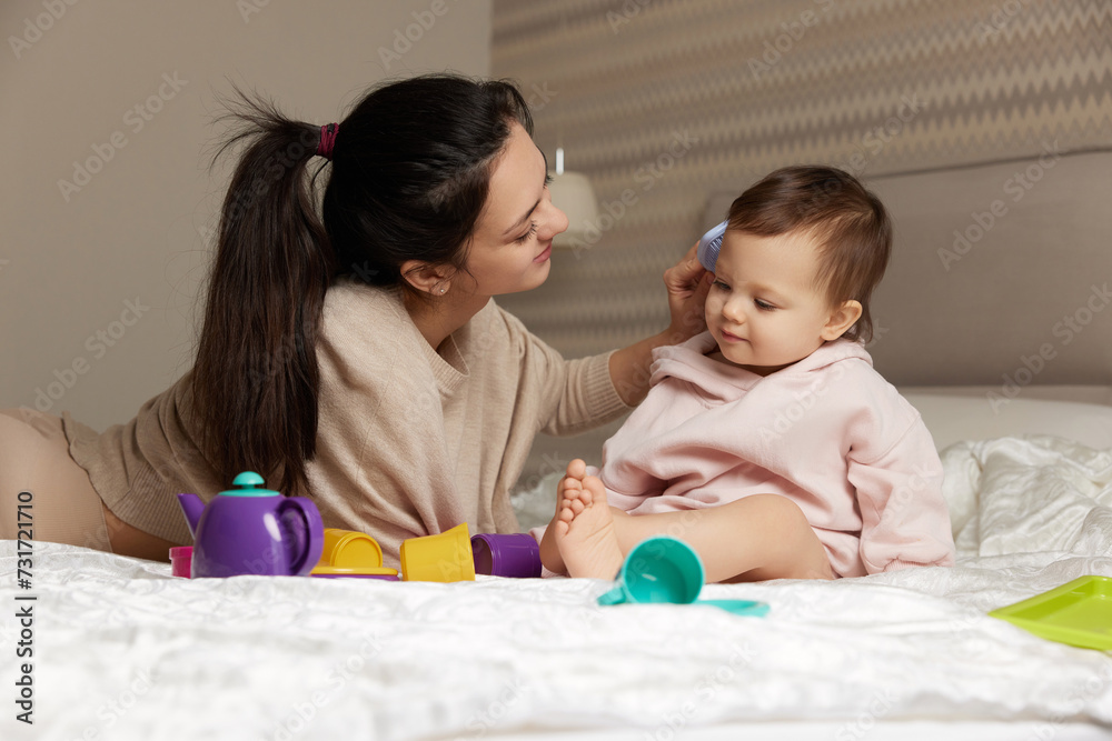Happy brunette mother is combing her cute little daughter hair on bed Happy brunette mother is combing her cute little daughter hair on bed