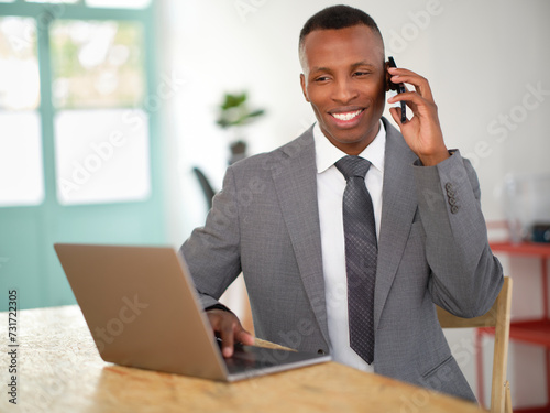 African-American business man on his laptop while talking on the phone. Black man smiling on his computer. Male in a corporate environment on his workstation. 