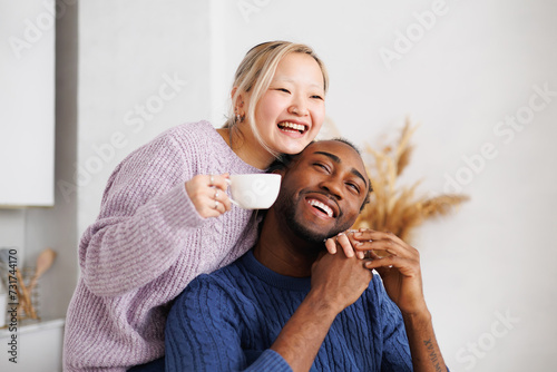 Smiling asian woman in sweater holding coffee cup and hugging african american boyfriend in kitchen
