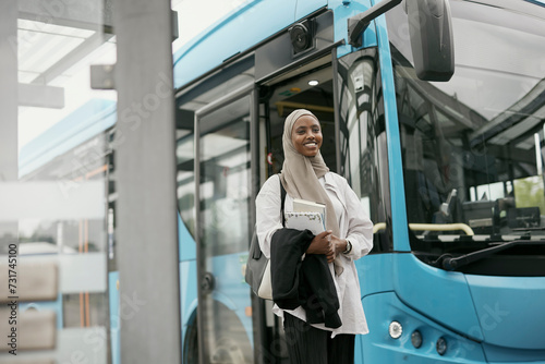 Smiling young woman wearing hijab standing in front of bus and looking at camera photo