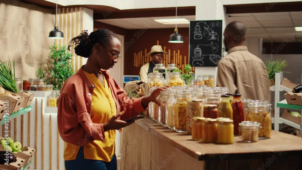 Customer checking shopping list on phone before choosing products, trying to buy organic locally grown food for low carbon footprint. African american woman looking at checklist notes. Camera B.