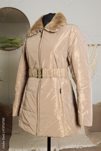 Women's winter jacket of medium length on a tailor's mannequin. Beige jacket with a fur collar and a buckle belt. Demonstration of clothes in an online store.