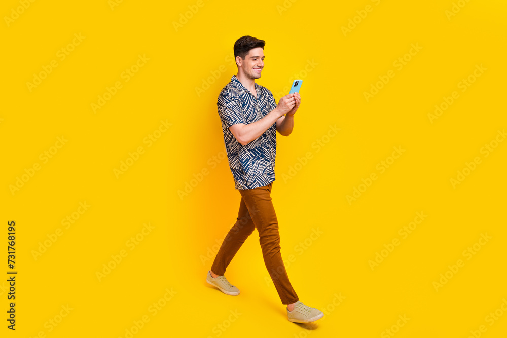 Fototapeta premium Full length photo of clever man dressed print shirt brown pants look at smartphone go empty space isolated on yellow color background