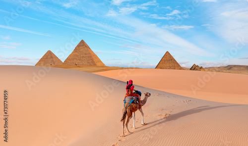 Camels in Giza Pyramid Complex - A woman in a red turban riding a camel across the thin sand dunes - Cairo, Egypt