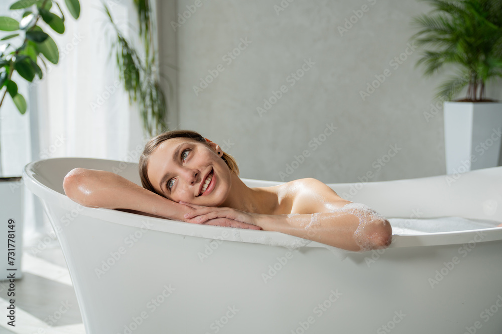 Obraz premium Pensive young woman, leaning on edge of white bathtub, while soaking in bathtub. Portrait of attractive, dreamy woman looking away, while taking bath at home, with blurred background. Spa concept.
