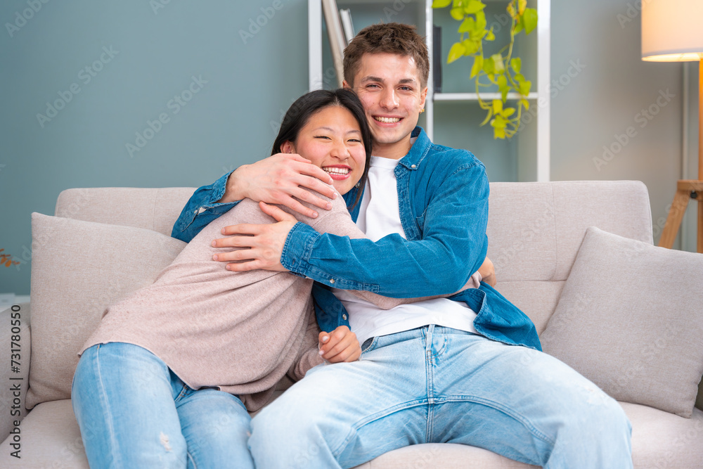 Close-up of a beautiful smiling young couple looking at the camera with tenderness and love while sitting in the living room of their home, young couple hugging inside Close-up of a beautiful smiling young couple looking at the camera with tenderness and love while sitting in the living room of their home, young couple hugging inside