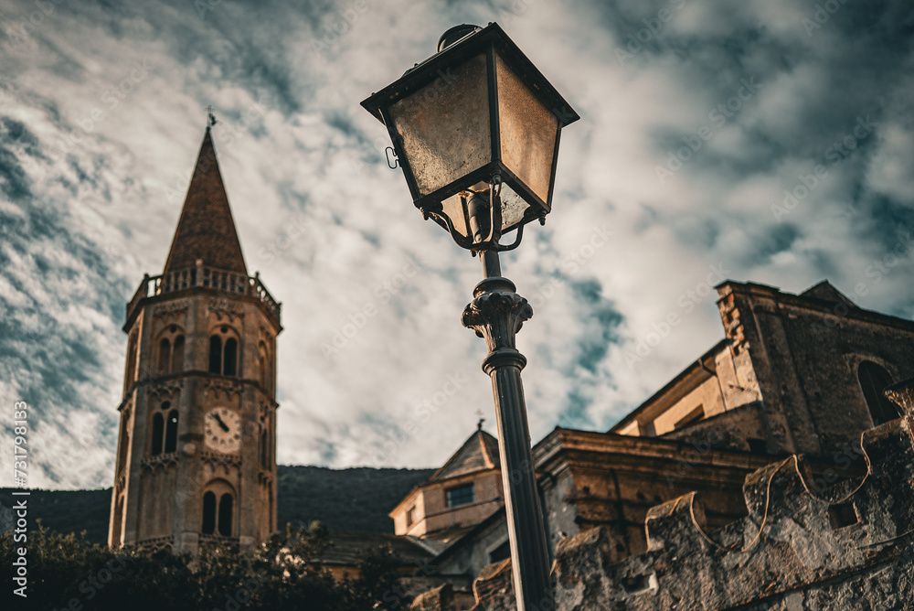 Village of Finalborgo, Finale Ligure, a medieval street lantern(in focus) with an old church(out of focus) on the background and a beautifull cloudy sky, sunset time, Italy Village of Finalborgo, Finale Ligure, a medieval street lantern(in focus) with an old church(out of focus) on the background and a beautifull cloudy sky, sunset time, Italy