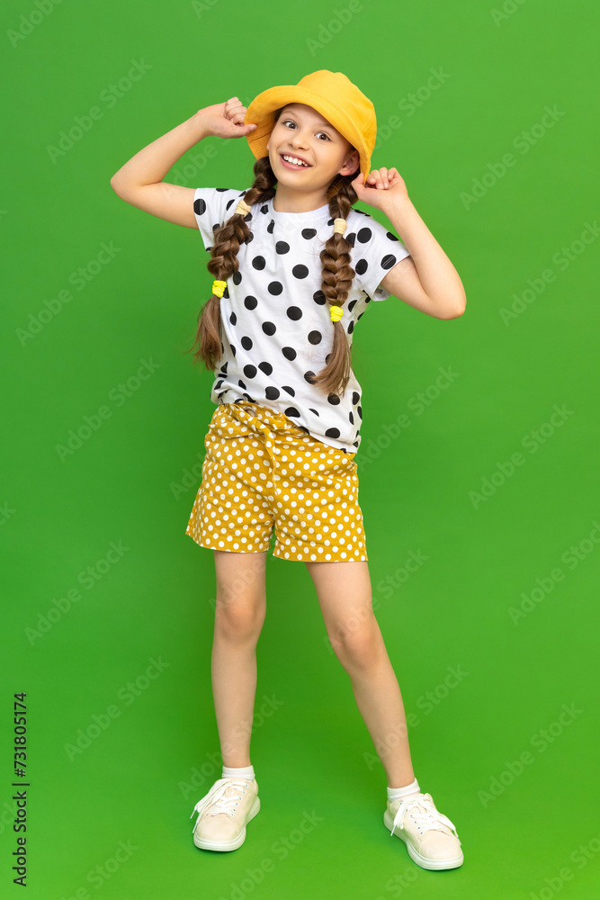 A full-length teenage girl dressed in a summer hat and shorts. A beautiful little lady is holding onto her hat and smiling broadly. Summer clothes for recreation. Green isolated background. A full-length teenage girl dressed in a summer hat and shorts. A beautiful little lady is holding onto her hat and smiling broadly. Summer clothes for recreation. Green isolated background.