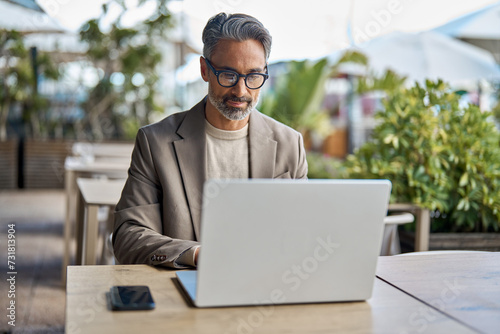 Busy middle aged business man executive ceo wearing suit and glasses sitting at outdoor table using laptop. Mature professional businessman manager hybrid working looking at computer outside office.