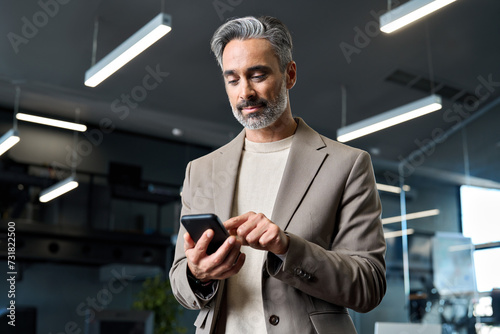 Busy mature older business man wearing suit using mobile cell phone at work. Middle aged businessman executive manager or entrepreneur holding smartphone looking at cellphone standing in office. photo