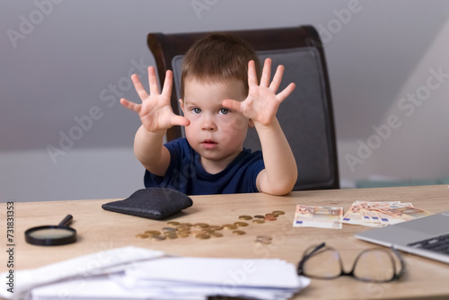 A boy in a blue T-shirt sits at a table with money, a magnifying glass and a wallet and shows a finger with open palms. Business concept. High quality photo