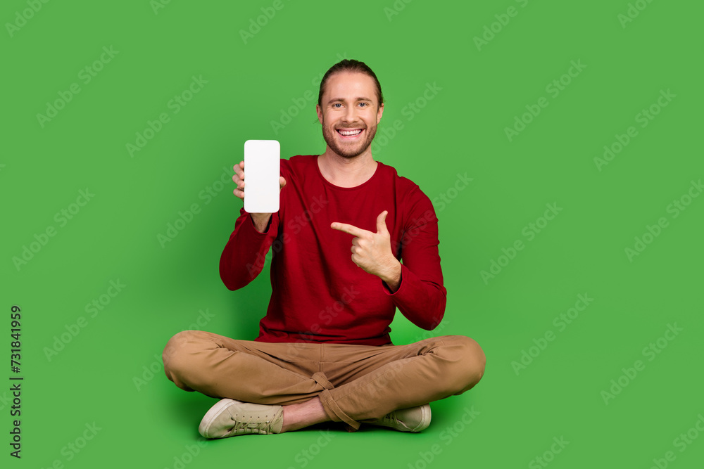 Full length photo of clever man dressed pullover sit indicating at smartphone screen empty space isolated on green color background Full length photo of clever man dressed pullover sit indicating at smartphone screen empty space isolated on green color background