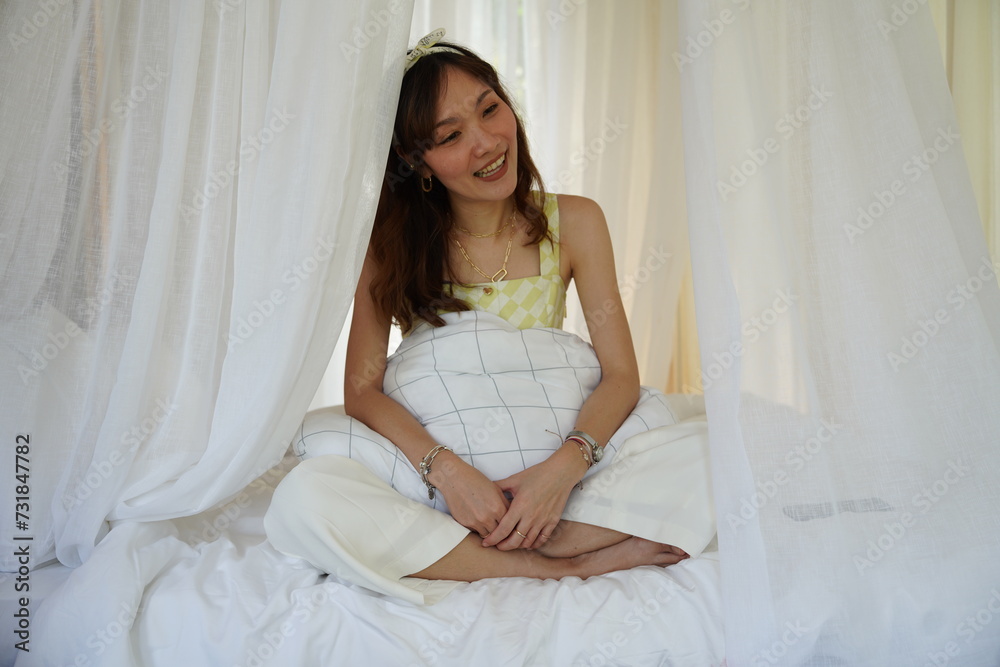 Asian woman wearing white pajamas wakes up early in the morning and sits by the window enjoying the view outside in her bedroom at a resort house.