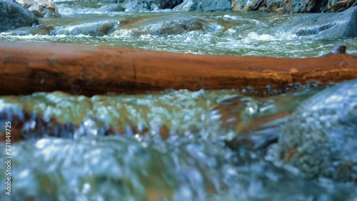 Wood blocks river. Water flowing through a river against a tree branch. Tree trunk in the middle of a river with water falling and rocks in the background. Extreme close up of mountain river. Slow mo photo