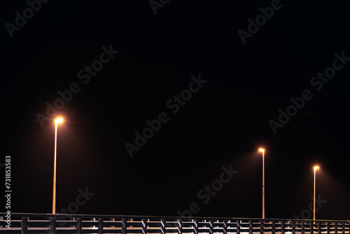 Street lampposts casts dim yellow warm along shore pier standing against pitch black canvas of night sky paints poetic picture of pier in stillness of night, serenity of solitary moments photo