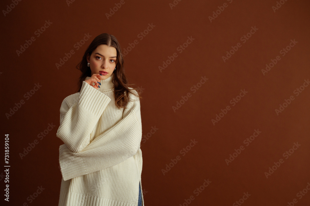 Fashionable confident brunette woman wearing oversized white knitted turtleneck sweater, posing on brown background. Studio fashion portrait. Copy, empty blank space for text