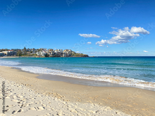 Beach with waves curling on the sand. Shore with sun and clouds. Village houses on a hill in the distance.