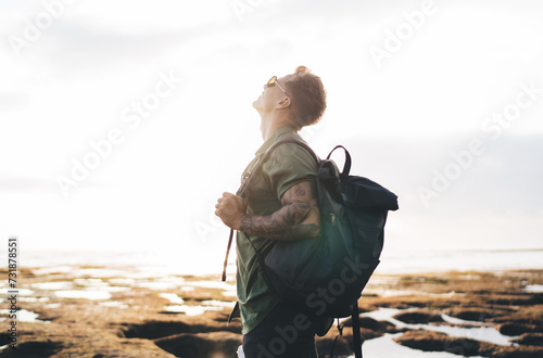 Carefree male traveler admiring view of sky near ocean shore with backpack