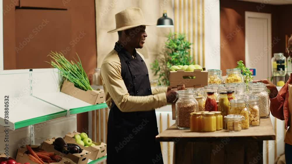 Local vendor placing crates with freshly harvested fruits on grocery store display, recommending organic produce to regular customer. African american farmer restocking all merchandise. Camera A. Local vendor placing crates with freshly harvested fruits on grocery store display, recommending organic produce to regular customer. African american farmer restocking all merchandise. Camera A.