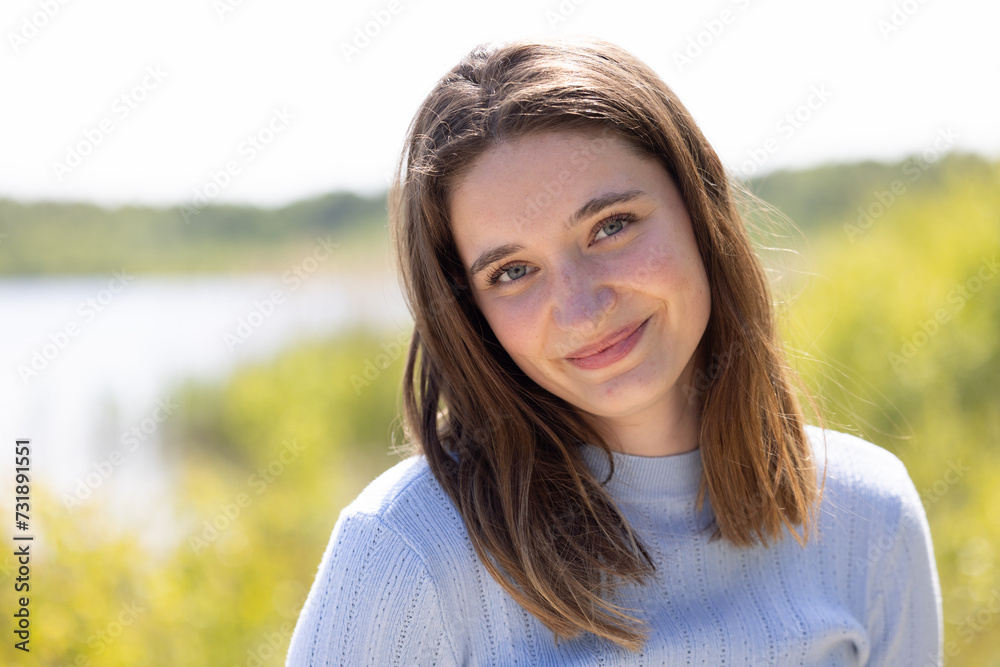 This image features a young woman with a soft, serene smile, standing by a lakeside. She is dressed in a light blue sweater, which adds a gentle touch to the overall warmth of the scene. The This image features a young woman with a soft, serene smile, standing by a lakeside. She is dressed in a light blue sweater, which adds a gentle touch to the overall warmth of the scene. The