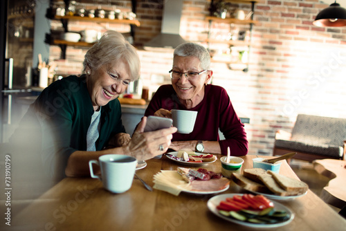 Two senior women eating breakfast with a smartphone in the kitchen