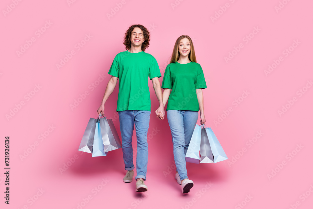 Full body length photo of two teen best friends walking holding arms shopping bags make purchases isolated on pink color background Full body length photo of two teen best friends walking holding arms shopping bags make purchases isolated on pink color background