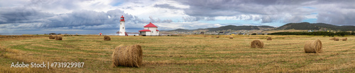 Wide panorama of the Anse a la Cabane lighthouse, the oldest working lighthouse on the Magdalen Islands, Gulf of Sait Lawrence, Canada. Rural setting with hay rolls in the foreground. © Rixie