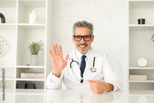 Photography A genial mature doctor in a white coat waves hello, inviting a sense of comfort and accessibility, seated in a well-organized clinic office that reflects a clean and efficient medical environment