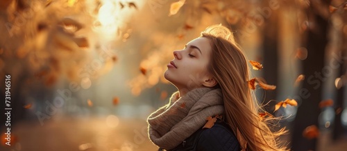 A happy blond woman, enjoying the heat, stands in a park with her eyes closed surrounded by falling leaves, experiencing nature's art and having fun.