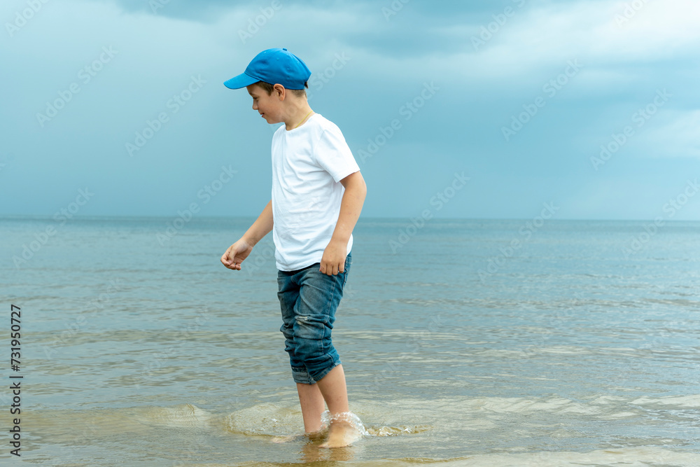 A young boy in a blue hat is standing in the water on the beach