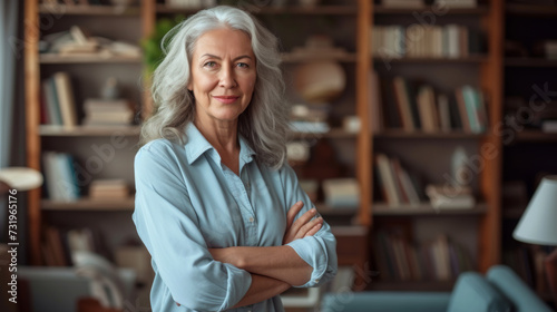 an elegant senior woman with grey hair, smiling at the camera, arms crossed, standing in a room with a bookshelf filled with books in the background