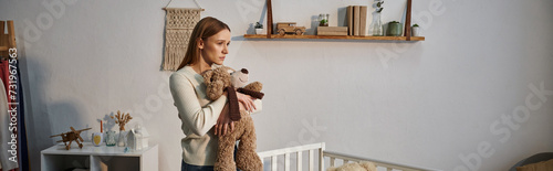 depressed young woman with soft toy standing near crib in dark nursery room at home, banner photo